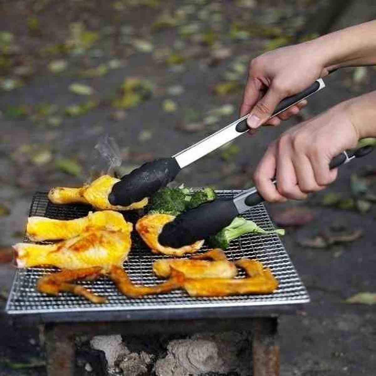 Pince à barbecue en acier inoxydable et silicone noir utilisée pour retourner du poulet et des brocolis sur une grille de barbecue en extérieur.