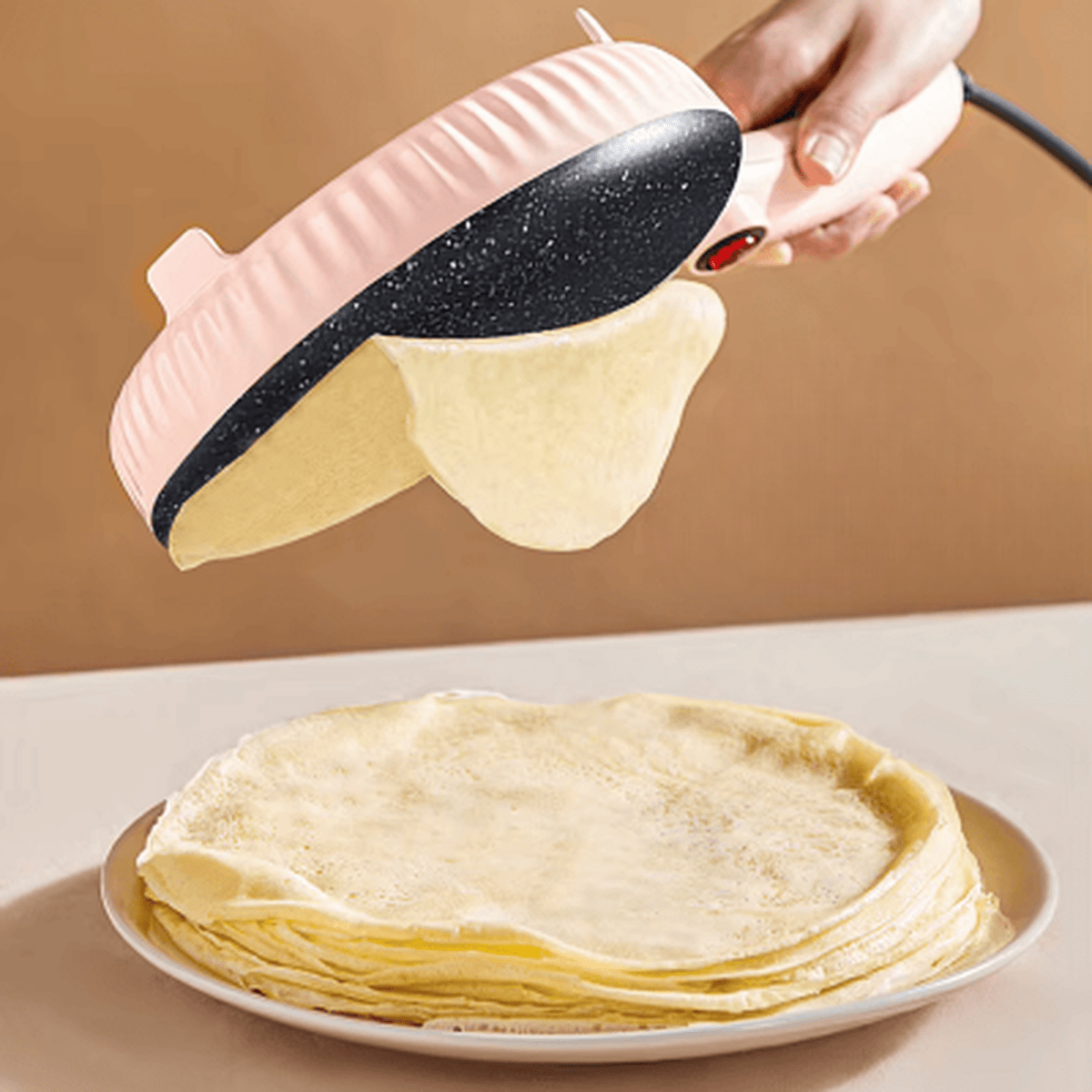 Démonstration de la poêle à crêpe électrique rose PancakeMaker créant une crêpe fine au-dessus d'une pile de crêpes dorées.